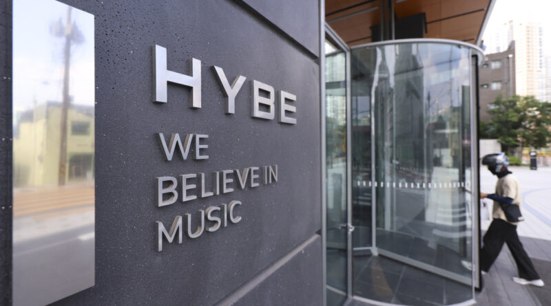 A trading floor showing the KOSPI index; BTS's comeback in 2026k;MediaXTrand ; The HYBE logo displayed at a press event; Investors monitoring K-pop stock charts.