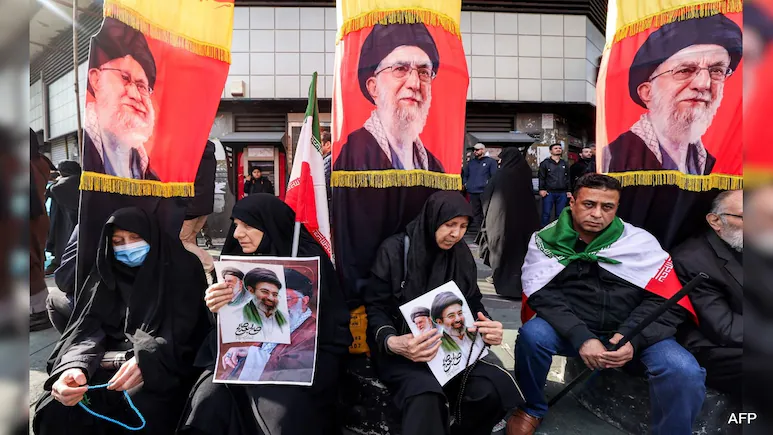 Smoke rising over Tehran skyline; US B-1 bombers in flight; Oil price ticker showing $119 surge.